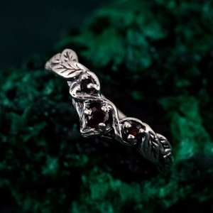 Può includere: Un anello d'argento con un design a foglie e viti, con tre piccole pietre preziose rosso scuro a taglio quadrato. L'anello ha un aspetto dettagliato e strutturato, con le foglie e le viti che avvolgono la fascia.