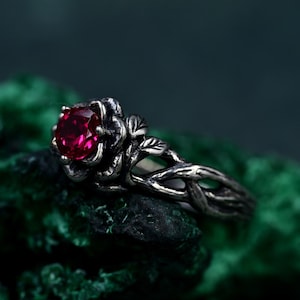 May include: A silver ring featuring a vibrant red gemstone set in a rose-shaped design. The ring's band has a detailed, vine-like pattern. The gemstone is the focal point, set against a dark green background.