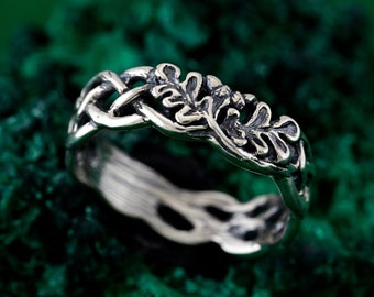 May include: A silver-toned ring featuring an intricate band design that combines a woven, braided pattern with detailed, raised oak leaf motifs. The metal has a darkened, oxidized finish in the recessed areas to highlight the texture.