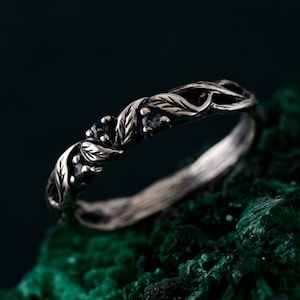 May include: A silver ring featuring a delicate design of intertwined leaves and small blue gemstones. The ring is set against a dark green background.