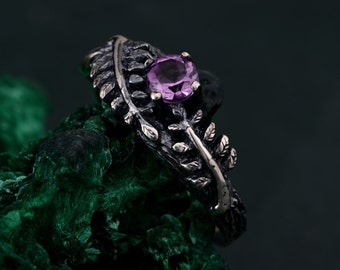 May include: A silver ring with a leaf design and a round, purple gemstone. The ring has intricate detailing on the band, resembling a delicate leaf pattern. The gemstone is set in a prong setting, adding a touch of elegance.