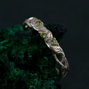 May include: A silver ring featuring a delicate, leafy design and three small green gemstones. The ring is set against a dark green background.