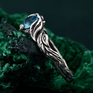 May include: A silver ring with a blue gemstone. The ring has a nature-inspired design, with the band and setting resembling intertwined branches. The blue gemstone is oval-shaped and sits prominently in the center, creating a striking contrast against the silver.