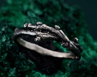 May include: A silver-toned ring with a branch-like design. The ring features a textured band and a detailed, twig-like top with small, bud-like accents. The ring is set against a dark green, textured background.