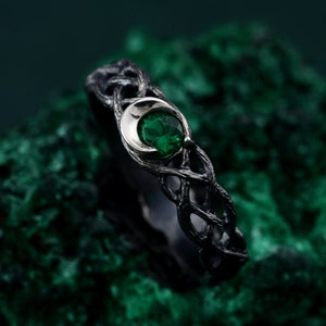 May include: A silver ring with a black oxidized finish and a green gemstone set in a crescent moon design. The ring is on a green background.
