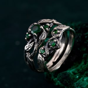 Può includere: Due anelli d'argento con un design ispirato alla natura. Gli anelli presentano intricati dettagli di foglie e viti, adornati da più pietre preziose verdi rotonde e vibranti. Gli anelli sono su uno sfondo verde scuro.