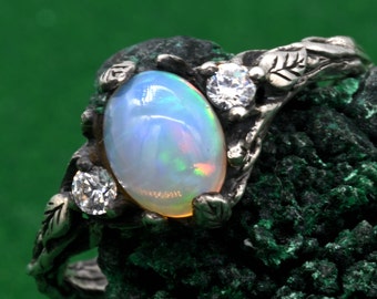May include: A silver ring featuring an oval opal gemstone with iridescent colors. The ring is accented with two clear, round gemstones and leaf-like details. The ring is set against a green background.