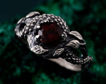 May include: A sterling silver ring featuring a large, faceted, round, deep red gemstone. The ring has an intricate, nature-inspired design with leaves and textured details. The gemstone is held in place by four prongs.