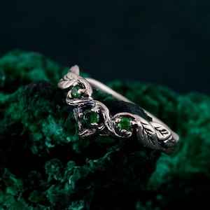 Può includere: Un anello d'argento con un design a foglie e vite, con tre pietre preziose verde smeraldo. La fascia dell'anello è delicatamente curva e le foglie presentano venature dettagliate. Sfondo verde scuro strutturato.