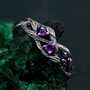 May include: A silver ring with a twisted branch design and three purple gemstones. The ring is set on a green background.