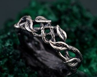 May include: A silver ring with a nature-inspired design. The ring features a detailed pattern of leaves and vines, creating an organic and textured appearance. The ring is set against a dark green background.