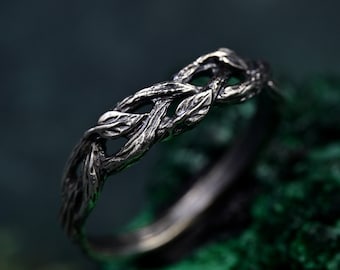 May include: A silver-toned ring with a nature-inspired design. The ring features a woven pattern resembling intertwined branches and leaves. The ring is set against a dark green background.