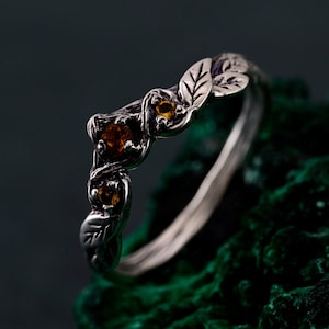 Op de afbeelding: Een zilveren ring met een delicaat bladontwerp en drie amberkleurige edelstenen. De ring ligt op een groene achtergrond.