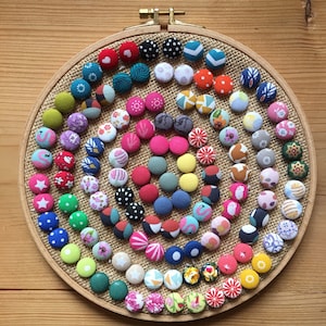 Peut inclure: Une collection de boutons recouverts de tissu coloré disposés en spirale sur un fond en toile de jute. Les boutons sont affichés sur un cercle de broderie en bois.