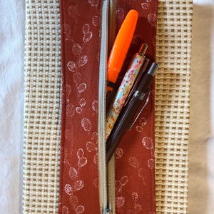 Puede incluir: Una libreta marrón y beige con un bolsillo con cremallera en la parte delantera. El bolsillo está forrado con una tela roja con un estampado floral blanco. Se pueden ver tres bolígrafos dentro del bolsillo, incluido un rotulador naranja.
