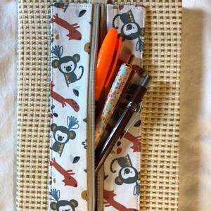 Puede incluir: Una libreta beige con un bolsillo con cremallera. El bolsillo está forrado con una tela blanca con un patrón de monos y jirafas rojas y naranjas. Se pueden ver tres bolígrafos en el bolsillo.
