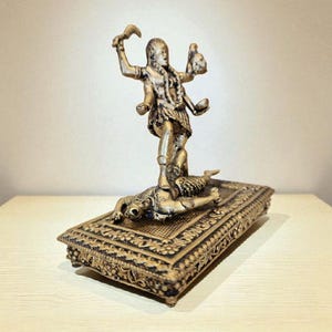 May include: A bronze-coloured statue of a multi-armed deity standing on a rectangular base. The base has intricate carvings and a figure lying on it. The deity holds objects in its hands, and the statue is set against a neutral background.