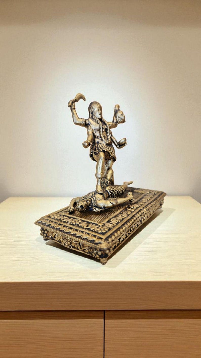 May include: A bronze-coloured statue of a multi-armed deity standing on a rectangular base. The base has intricate carvings and a figure lying on it. The deity holds objects in its hands, and the statue is set against a neutral background.