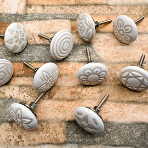 Peut inclure: Ensemble de onze boutons de meubles en céramique blanche avec divers motifs floraux et géométriques en relief. Chaque bouton a une tige en métal argenté. Les boutons sont disposés sur une surface en pierre texturée multicolore.