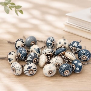May include: A selection of decorative ceramic cabinet knobs in various designs and colours, including white, navy blue, and cream. The knobs feature floral and geometric patterns with brass hardware. They are arranged on a light wood surface.