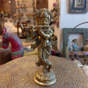 May include: A gold-colored metal statue of a Hindu deity playing a flute. The statue is standing on a round, ornate base with a floral design.