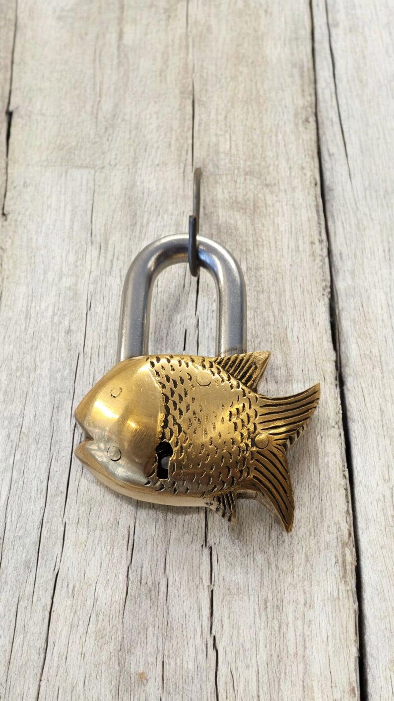May include: A decorative padlock with a fish-shaped brass body and a silver-colored shackle. The fish has detailed scales and fins. The lock is hanging against a weathered, light-colored wooden surface.