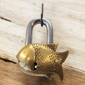 May include: A decorative padlock with a brass fish design. The fish has detailed scales and fins, with a silver-colored shackle. The padlock is hanging against a light brown wooden surface.