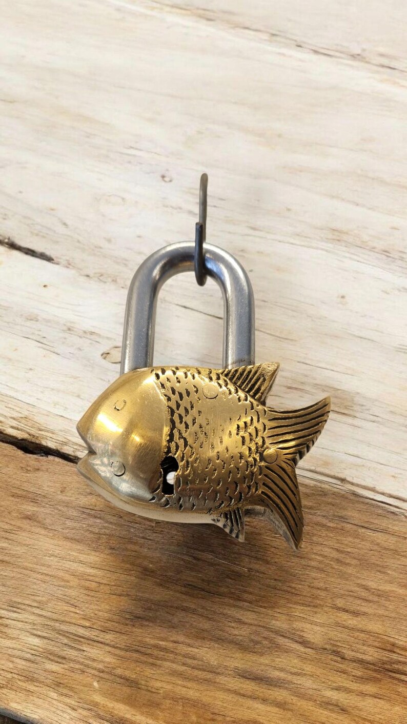 May include: A decorative padlock with a brass fish design. The fish has detailed scales and fins, with a silver-colored shackle. The padlock is hanging against a light brown wooden surface.