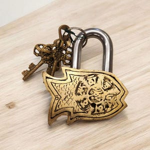 May include: A decorative padlock with a fish-shaped design in gold. The lock has a silver shackle and is accompanied by two ornate keys. The keys and lock are made of metal and rest on a light-colored wooden surface.