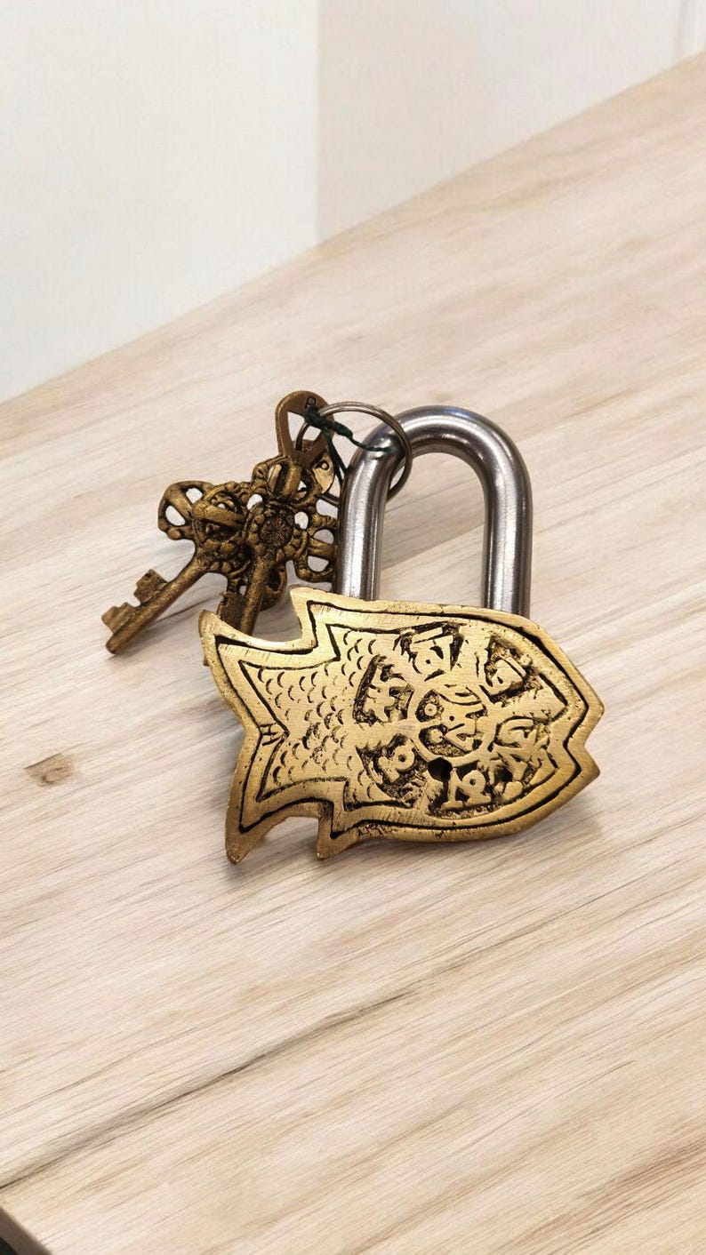 May include: A decorative padlock with a fish-shaped design in gold. The lock has a silver shackle and is accompanied by two ornate keys. The keys and lock are made of metal and rest on a light-colored wooden surface.