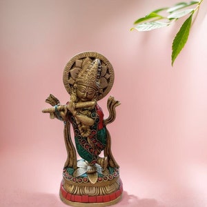 May include: A gold-colored statue of Krishna playing a flute, with a green and red patterned robe and a halo behind his head. The statue is standing on a lotus-shaped base with red and gold accents.