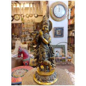 May include: A gold-colored metal statue of Krishna playing a flute. The statue is standing on a round base with intricate details. The statue is surrounded by a variety of other decorative items.