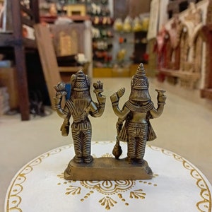 May include: A brass figurine of two deities standing back to back. The figurine is on a white circular base with gold painted designs.