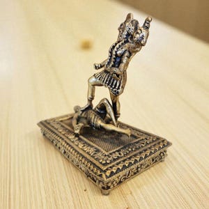 May include: A bronze-coloured figurine showing a figure standing over another on a rectangular base. The standing figure is in a dynamic pose, with one leg raised. The base has intricate patterns. The overall theme is historical or mythological.