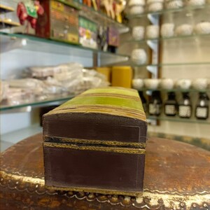 Puede incluir: Una peque&ntilde;a caja rectangular de madera con una tapa a rayas verde y marr&oacute;n. La caja tiene un borde dorado alrededor de los bordes y una base marr&oacute;n.