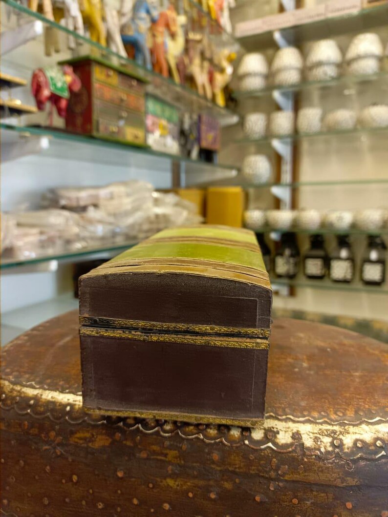 Puede incluir: Una peque&ntilde;a caja rectangular de madera con una tapa a rayas verde y marr&oacute;n. La caja tiene un borde dorado alrededor de los bordes y una base marr&oacute;n.