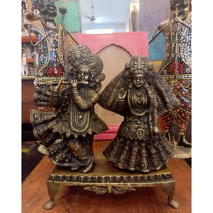 May include: A gold-colored metal statue of a couple, likely representing Krishna and Radha, standing on a decorative base with intricate details. The couple is depicted in traditional Indian attire, with the male figure holding a flute.