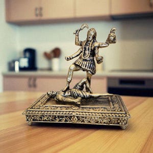May include: A detailed brass statue of a deity, standing on a figure, holding a sword and a severed head. The statue is on a rectangular, ornate base with intricate designs. The background shows a kitchen setting.
