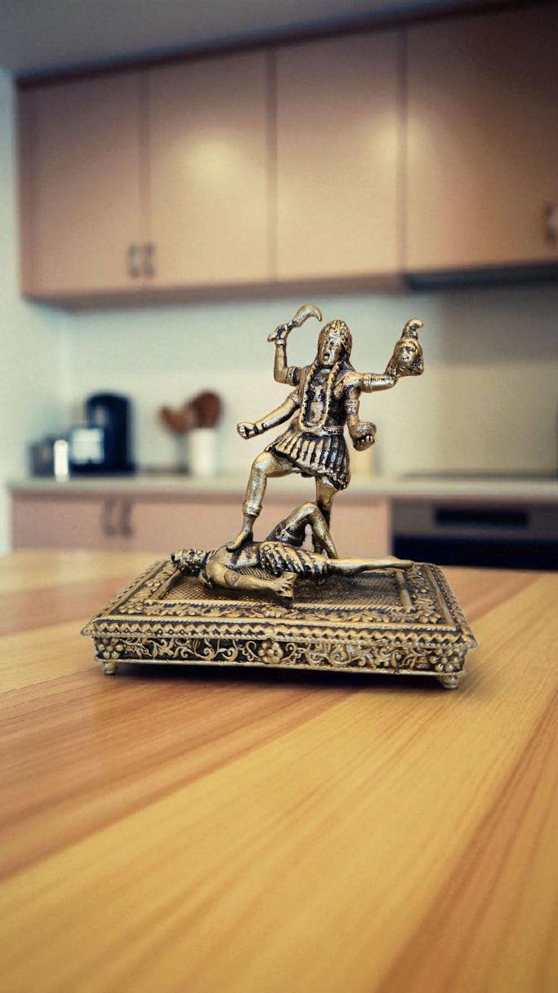 May include: A detailed brass statue of a deity, standing on a figure, holding a sword and a severed head. The statue is on a rectangular, ornate base with intricate designs. The background shows a kitchen setting.