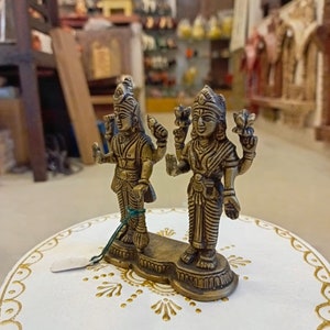 May include: A pair of gold-colored metal figurines of Hindu deities, standing on a white base with gold accents. The figurines are intricately detailed and have a traditional Indian style.