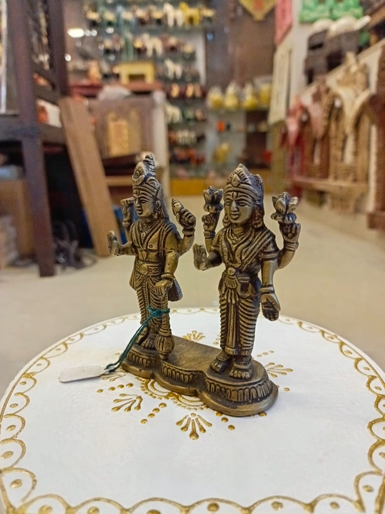 May include: A pair of gold-colored metal figurines of Hindu deities, standing on a white base with gold accents. The figurines are intricately detailed and have a traditional Indian style.