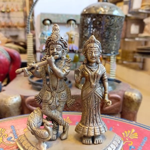 May include: A pair of gold-toned metal statues of Hindu deities, Radha and Krishna, standing on a decorative base. Krishna is playing a flute and Radha is holding a flower. The statues are intricately detailed and have a traditional Indian design.