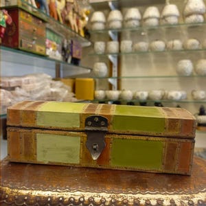 Puede incluir: Una peque&ntilde;a caja rectangular de madera con tapa abatible. La caja est&aacute; decorada con un mosaico de cuero verde, marr&oacute;n y naranja. La caja tiene un cierre de metal y un ribete de metal decorativo.