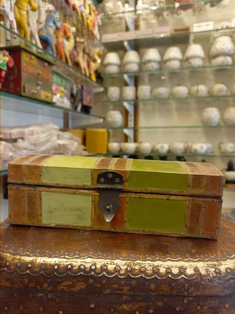 Puede incluir: Una peque&ntilde;a caja rectangular de madera con tapa abatible. La caja est&aacute; decorada con un mosaico de cuero verde, marr&oacute;n y naranja. La caja tiene un cierre de metal y un ribete de metal decorativo.