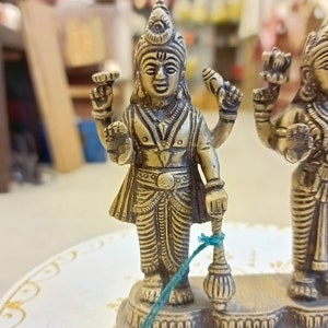 May include: A gold-colored metal statue of three Hindu deities. The central deity is holding a staff and has a third eye on their forehead. The statue is on a white surface with gold accents.