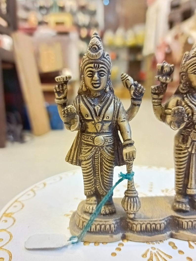 May include: A gold-colored metal statue of three Hindu deities. The central deity is holding a staff and has a third eye on their forehead. The statue is on a white surface with gold accents.