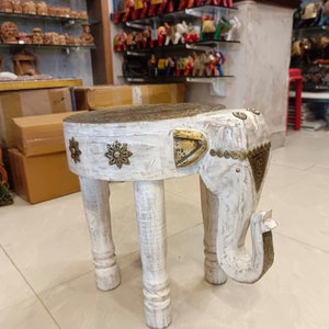 May include: White wooden elephant stool with gold accents and a carved design. The stool has three legs and a round top.