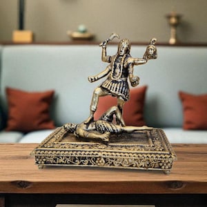 May include: A bronze-coloured statue of a multi-armed figure standing on a figure lying down. The statue is on a decorative base with intricate patterns. The figure holds a sword and a severed head.