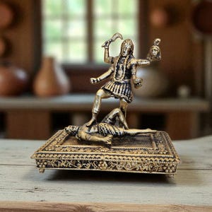 May include: A gold-coloured statue of a deity standing on a figure, holding a sword and a severed head. The statue is on a decorative rectangular base with ornate detailing. The background is blurred.