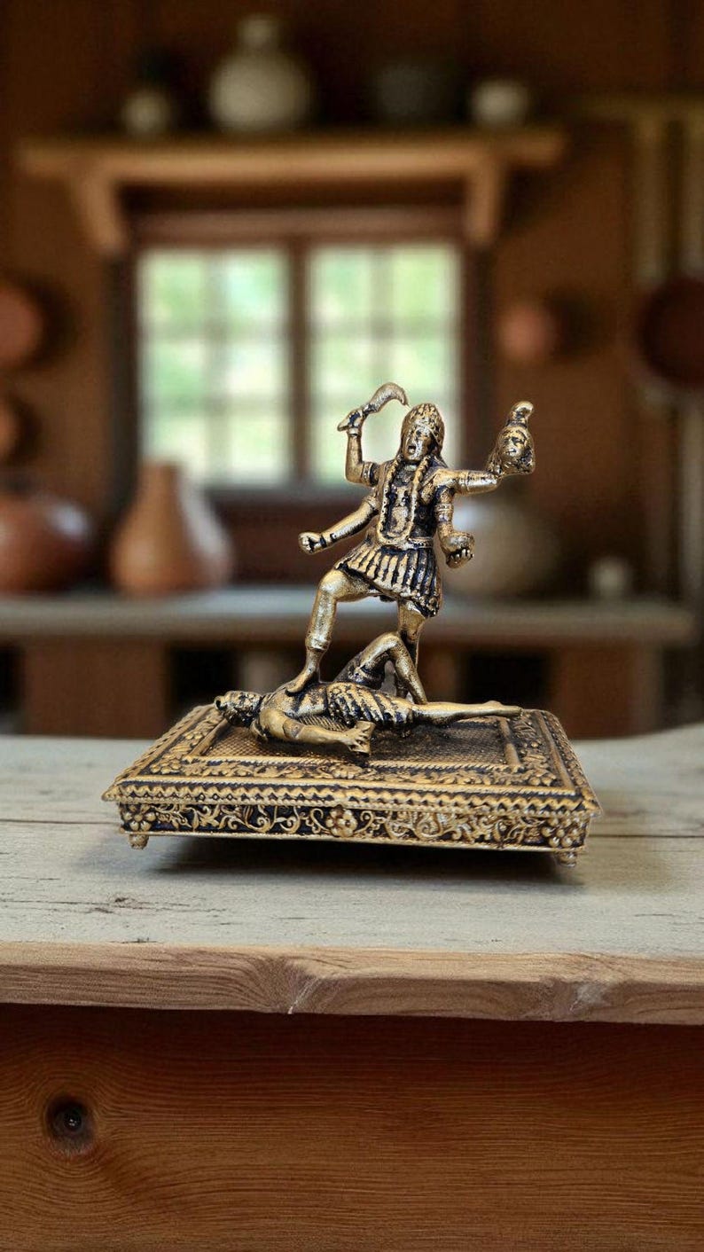 May include: A gold-coloured statue of a deity standing on a figure, holding a sword and a severed head. The statue is on a decorative rectangular base with ornate detailing. The background is blurred.